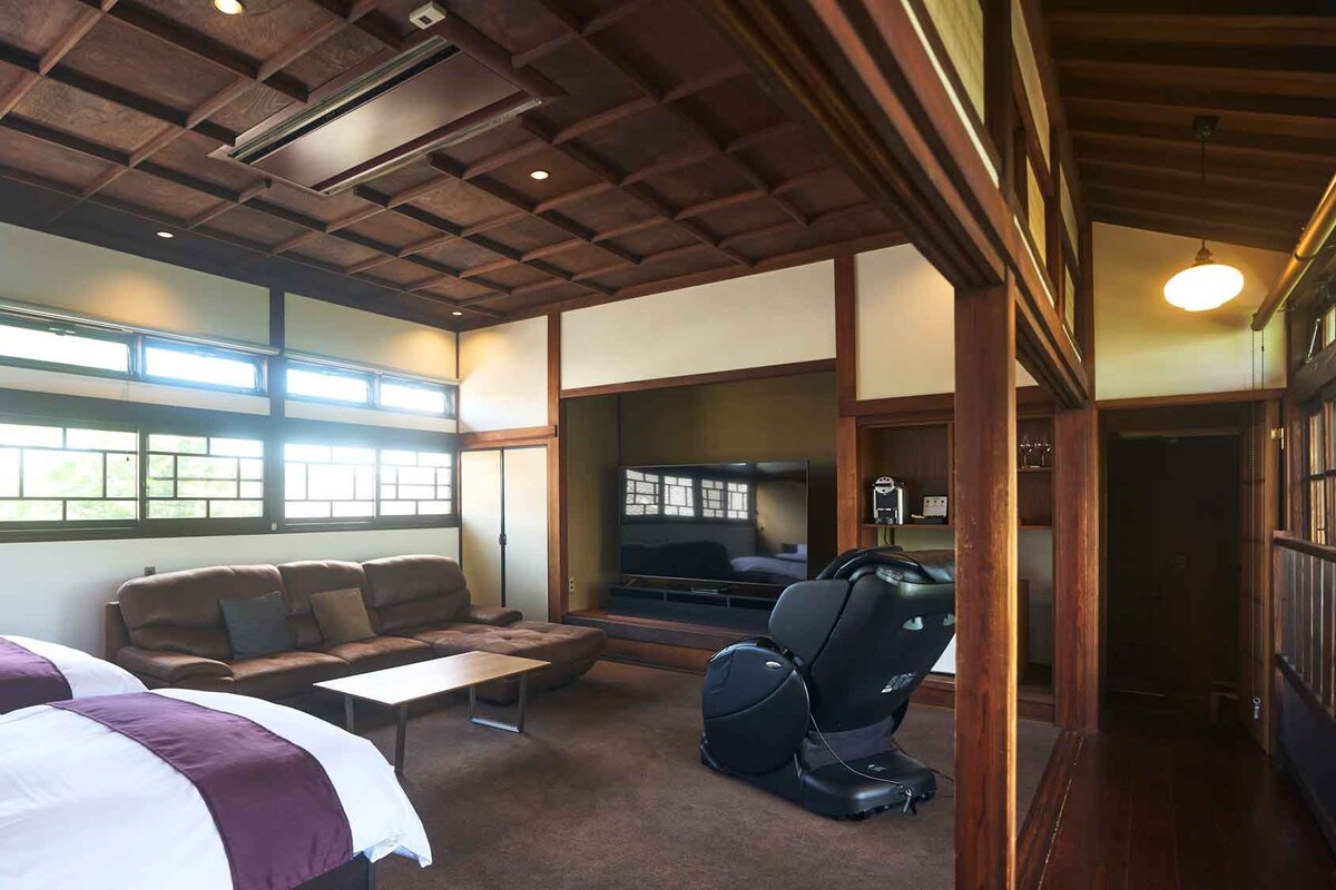 A hotel room with a bed, a sofa, a massage chair, a TV, and a large window.