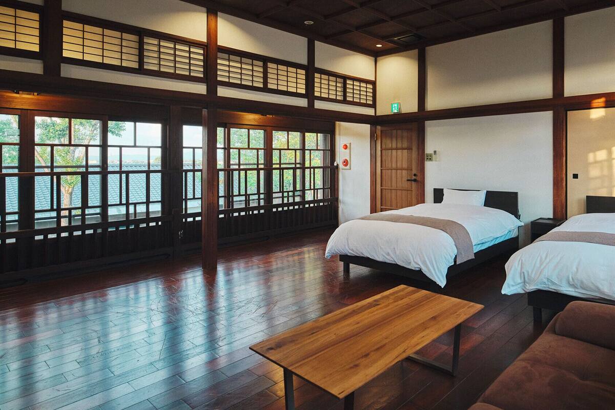 A traditional Japanese-style room with tatami flooring, two beds, a wooden table, and large windows.