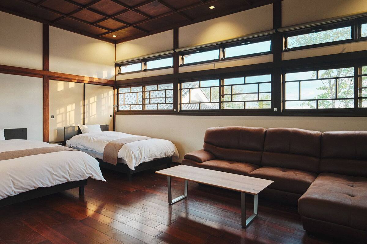 A hotel room with two beds, a sofa, a small table, and large windows.