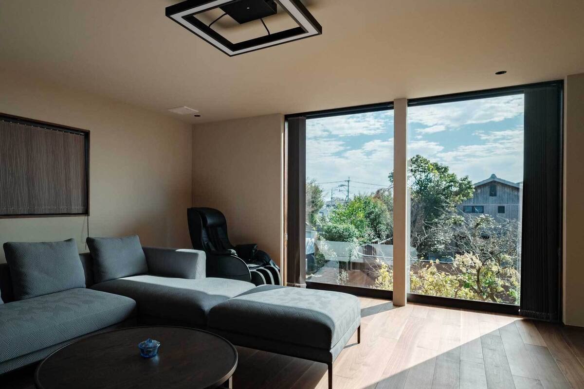 A modern living room with a sectional sofa, a massage chair, and large windows offering a view of the outside.
