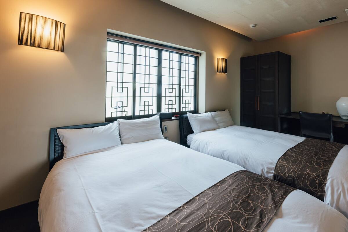 A hotel room with two beds, a window with a geometric pattern, and a desk with a lamp.