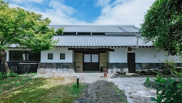 A traditional Japanese-style house with a tiled roof, a stone pathway leading to the entrance, and a small garden area with trees and plants.
