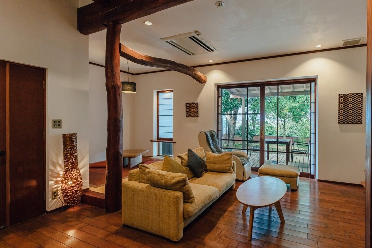 A spacious living area with a sofa, ottoman, and a round coffee table. There is a large window providing a view of the outdoors.
