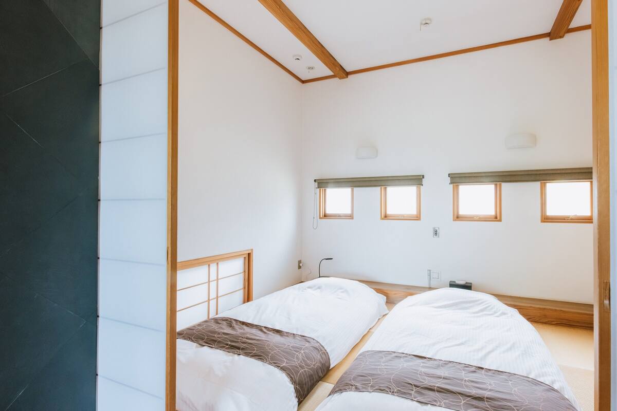 A minimalist room with two beds, a window, and a wooden ceiling structure.