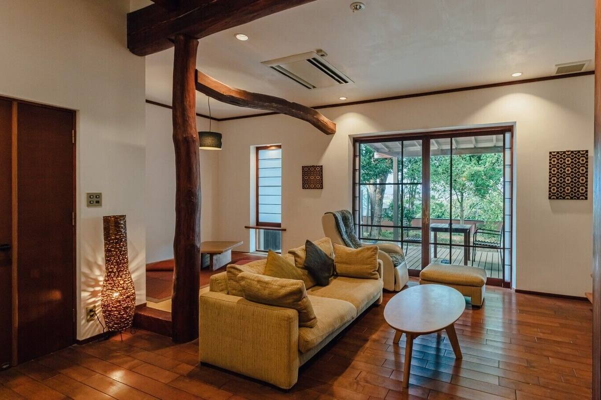 A spacious living area with a sofa, ottoman, and a round coffee table. There is a large window providing a view of the outdoors.