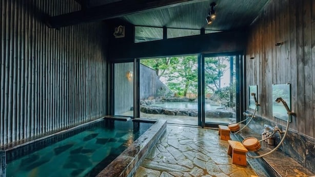 A spa area with a stone hot tub, wooden walls, and a view of a natural landscape. - Fukahoritei (Kurume)