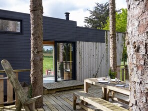 A wooden deck with a table and benches, a wooden chair, and a glass door leading to a garden. - La Cafoutche Tiny – 5 Guests with Private Terrace, Shared Garden, and Wi-Fi (Gommenec'h)