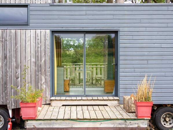 A modern building with a sliding glass door leading to an outdoor deck with a wooden railing. - La Cafoutche Tiny – 5 Guests with Private Terrace, Shared Garden, and Wi-Fi (Gommenec'h)