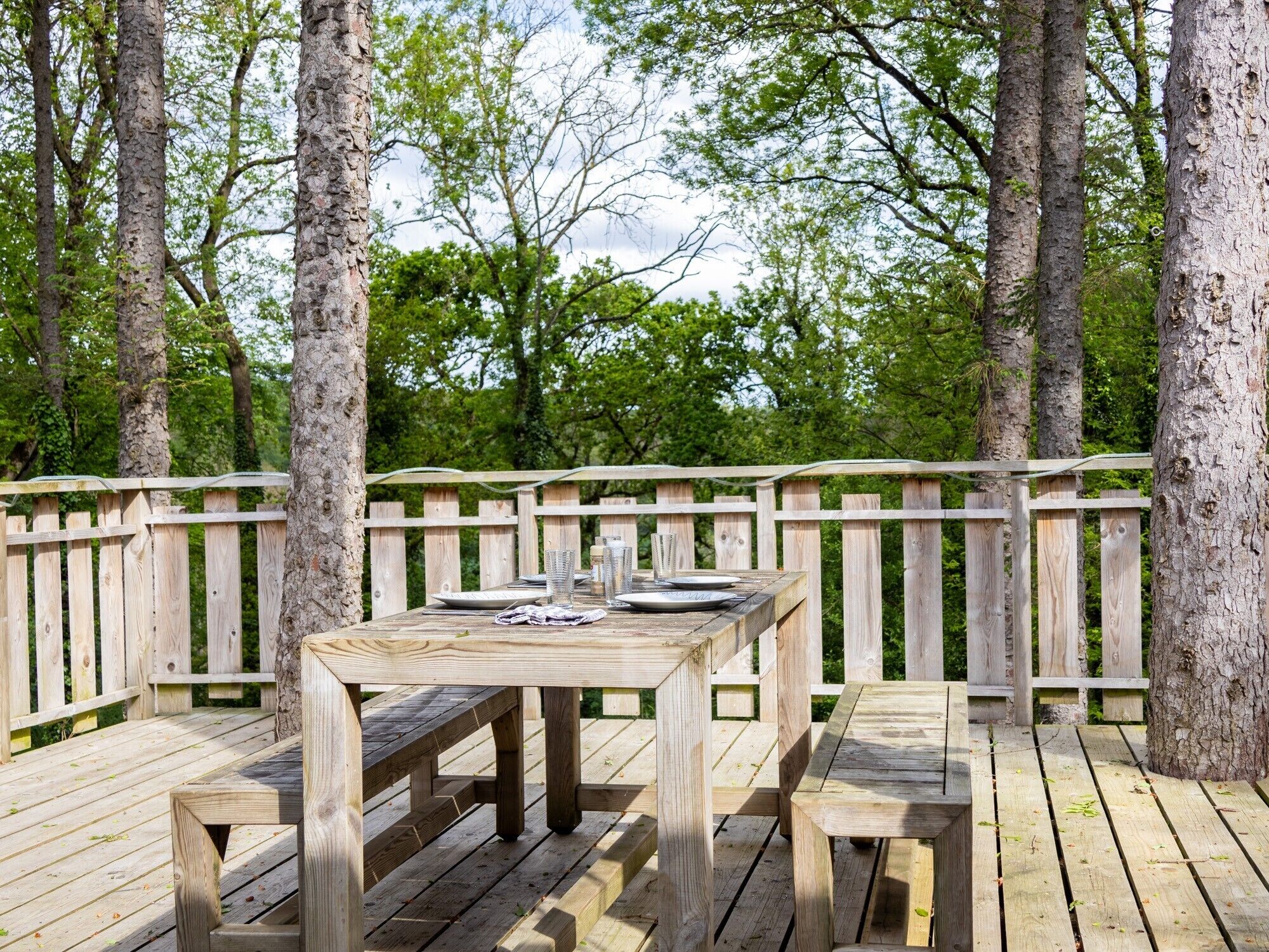 Une terrasse en bois avec une table et des bancs, entourée d’arbres.