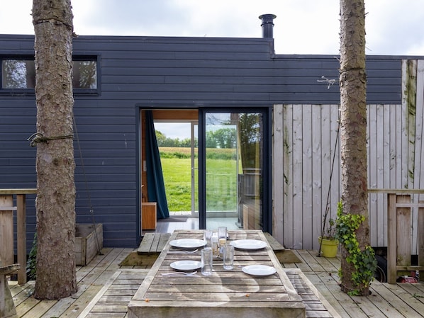 Une terrasse en bois avec une table dressée pour quatre personnes, une porte-fenêtre coulissante et une vue sur un champ verdoyant.