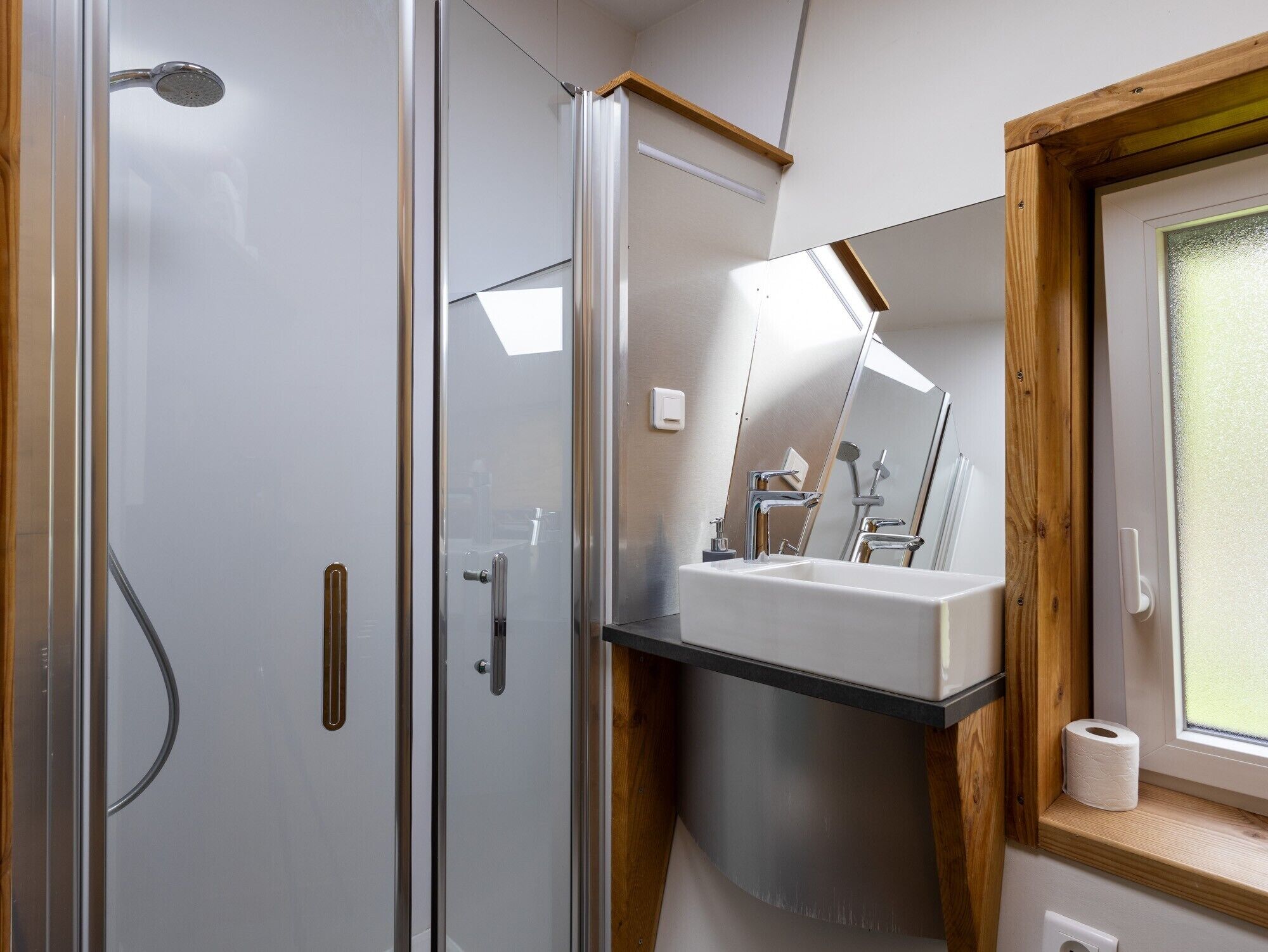 Un baño moderno con cabina de ducha de vidrio, un lavabo blanco y un marco de ventana de madera.
