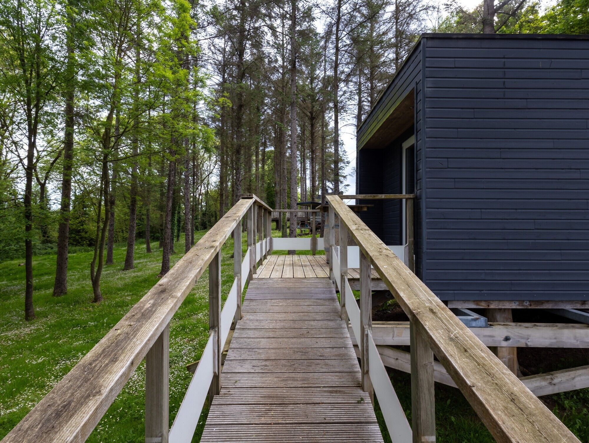 Une terrasse en bois mène à un chalet aux couleurs sombres, situé dans une zone boisée.
