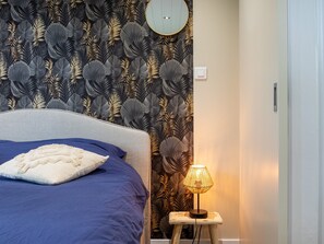 A bedroom with a bed, a nightstand with a lamp, a wall-mounted mirror, and a patterned wallpaper.