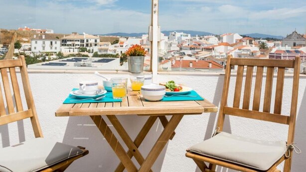Una mesa de madera con vistas a un paisaje urbano, dos sillas de madera y un cubo de metal con flores.