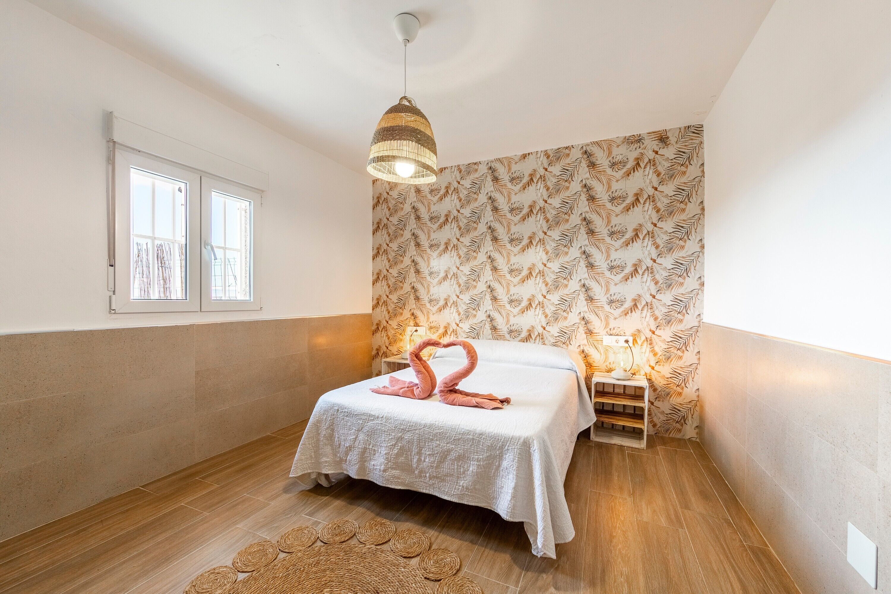 Un dormitorio con cama, lámpara fijada a la pared, ventana y papel tapiz estampado.