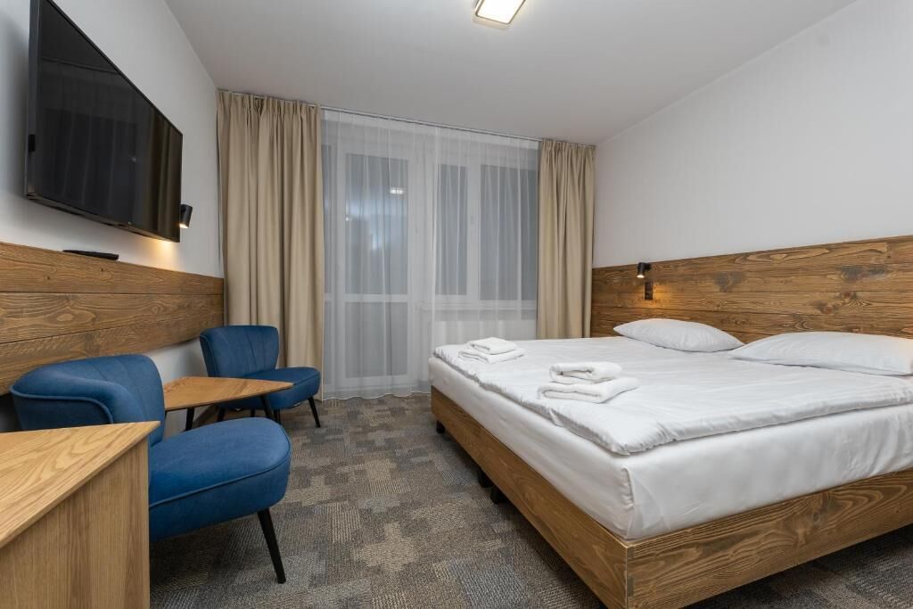 Habitación de hotel con cabecera de madera, cama con ropa blanca, una silla azul, una mesita de madera, televisor de pantalla plana montado en la pared y una ventana con cortinas translúcidas.
