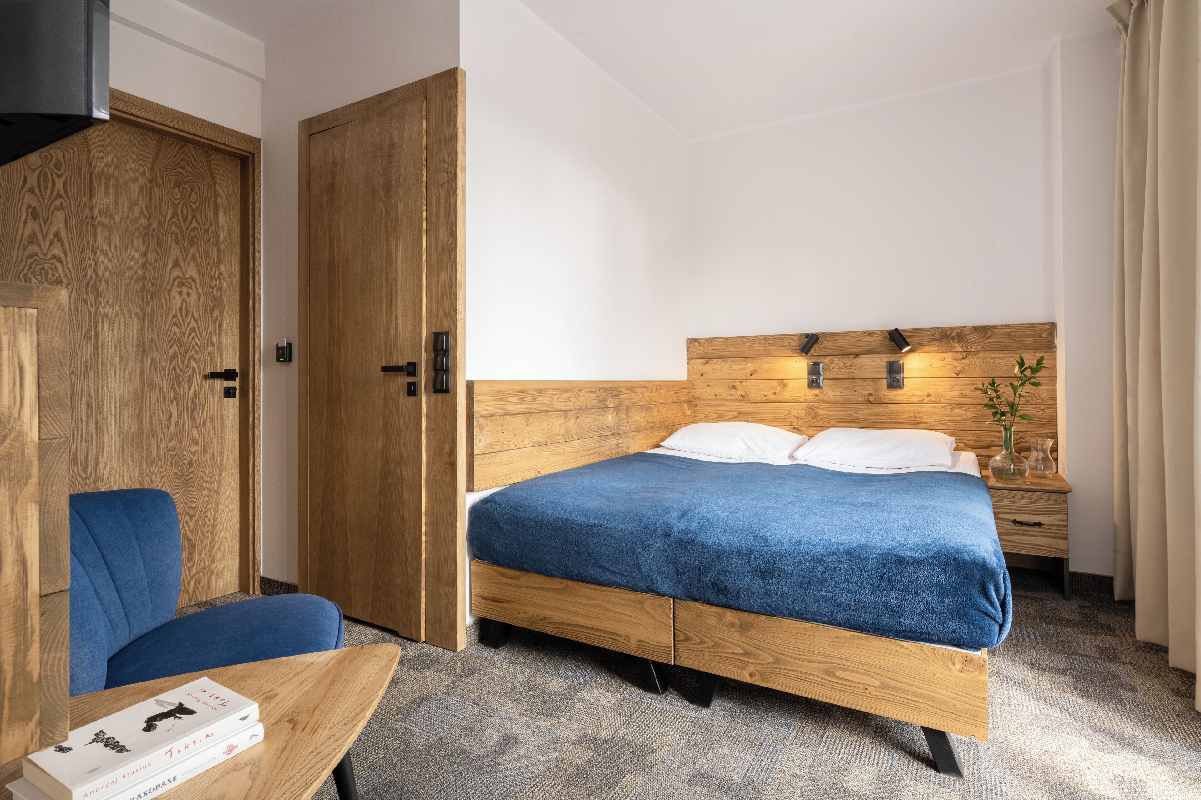 A hotel room with a wooden bed, a blue chair, a wooden wardrobe, and a small table with books.