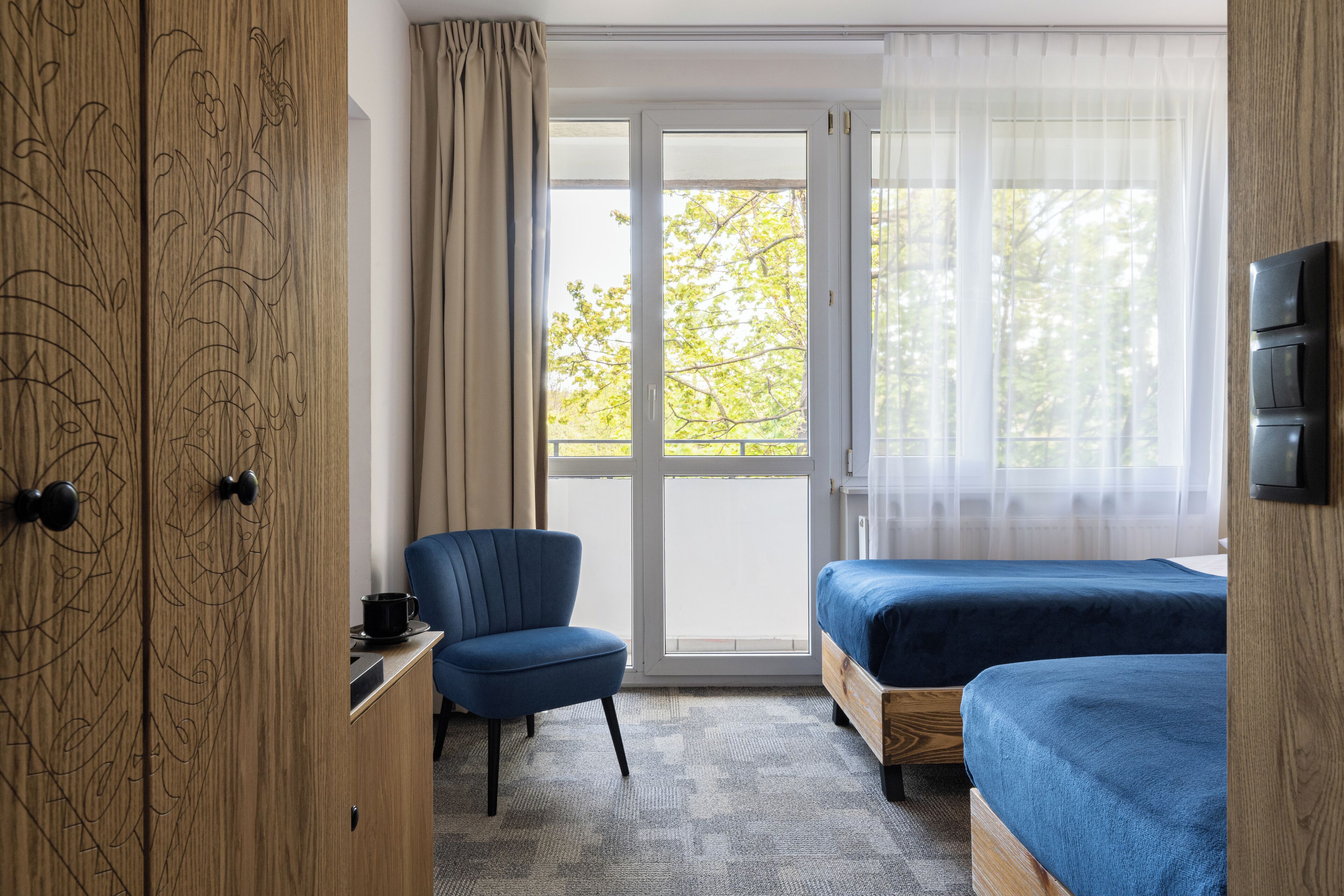 A room with a wooden wardrobe, a blue chair, a bed with a blue bedspread, and a large window with a view of trees.