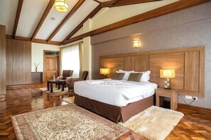 A spacious bedroom with a large bed, wooden headboard, two bedside lamps, a nightstand, a wooden wardrobe, a chair, a small table, and a patterned rug. - Gem Suites Hotel & Luxury Serviced Apartments (Nairobi)