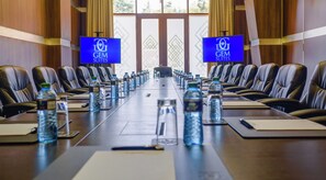 Un salón de conferencias con una larga mesa, varias sillas, botellas de agua y dos pantallas mostrando el logotipo de 'GEM SUITES'.