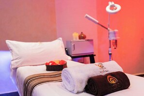 Una habitación de hotel con una cama, almohadas, una mesita con velas y un sillón de masaje.