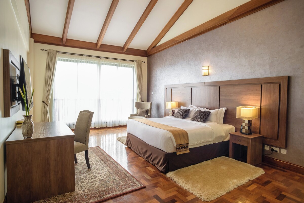 Habitación de hotel con una cama grande, un escritorio, una silla, una mesita de noche, una lámpara y vistas al exterior a través de una ventana.