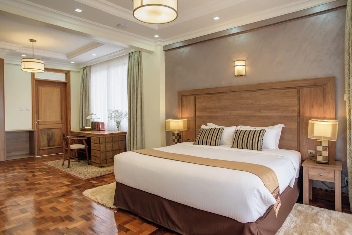 Habitación de hotel con una cama grande, cabecera de madera, dos lámparas de noche, un escritorio con silla y vistas al exterior por la ventana.