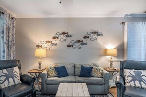 Un salon avec un canapé gris, deux fauteuils, une table basse et des œuvres d’art décoratives accrochées au mur.