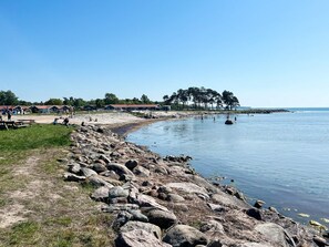 A lakeside area with a rocky shoreline, picnic tables, and a row of cabins in the background. - 4 Person Holiday Home in Farjestaden (Farjestaden)
