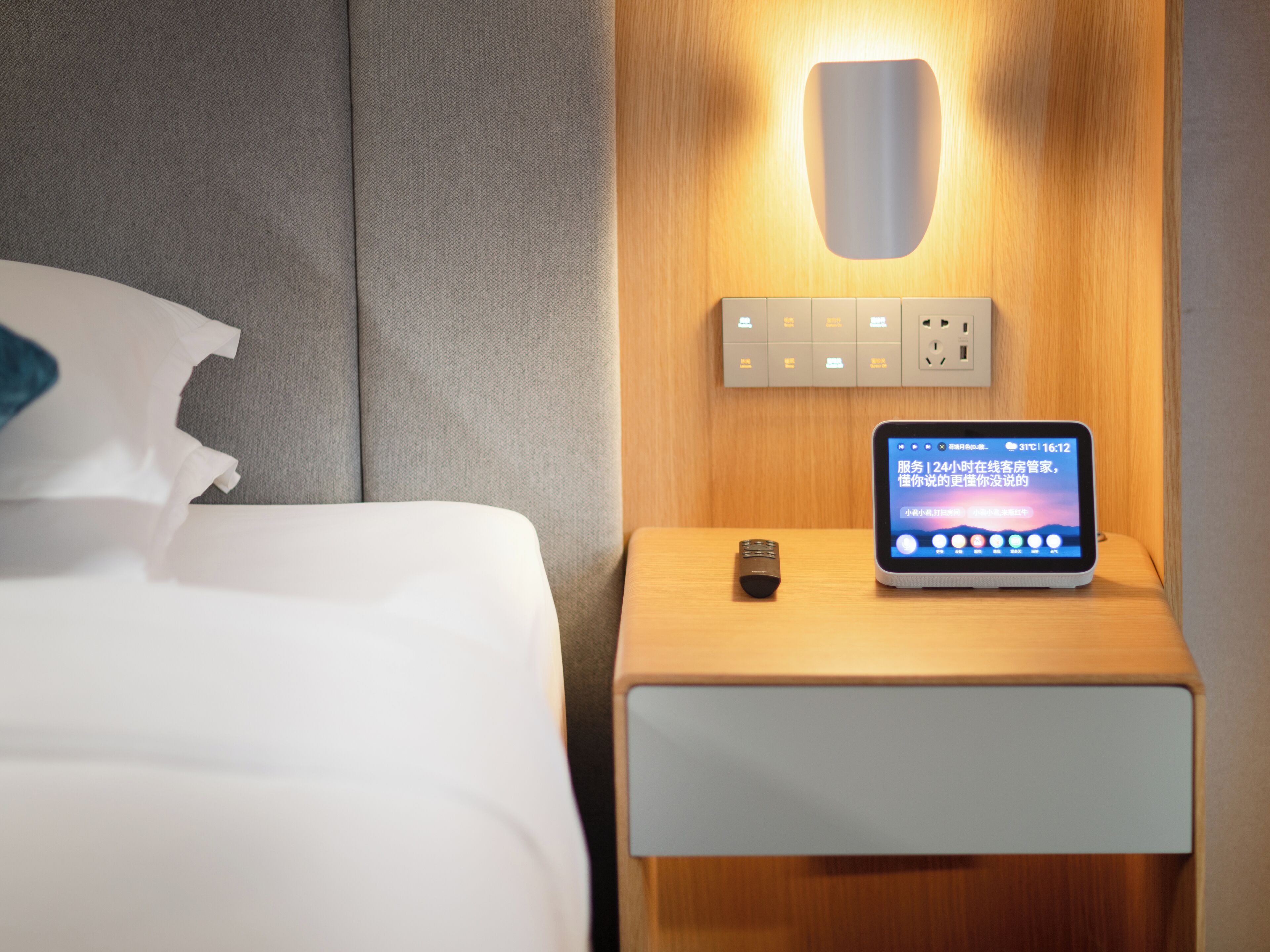 Een net opgemaakt bed met witte lakens en een blauw kussen, een nachtkastje met een smartphone waarop een bericht staat, en een wandlamp.