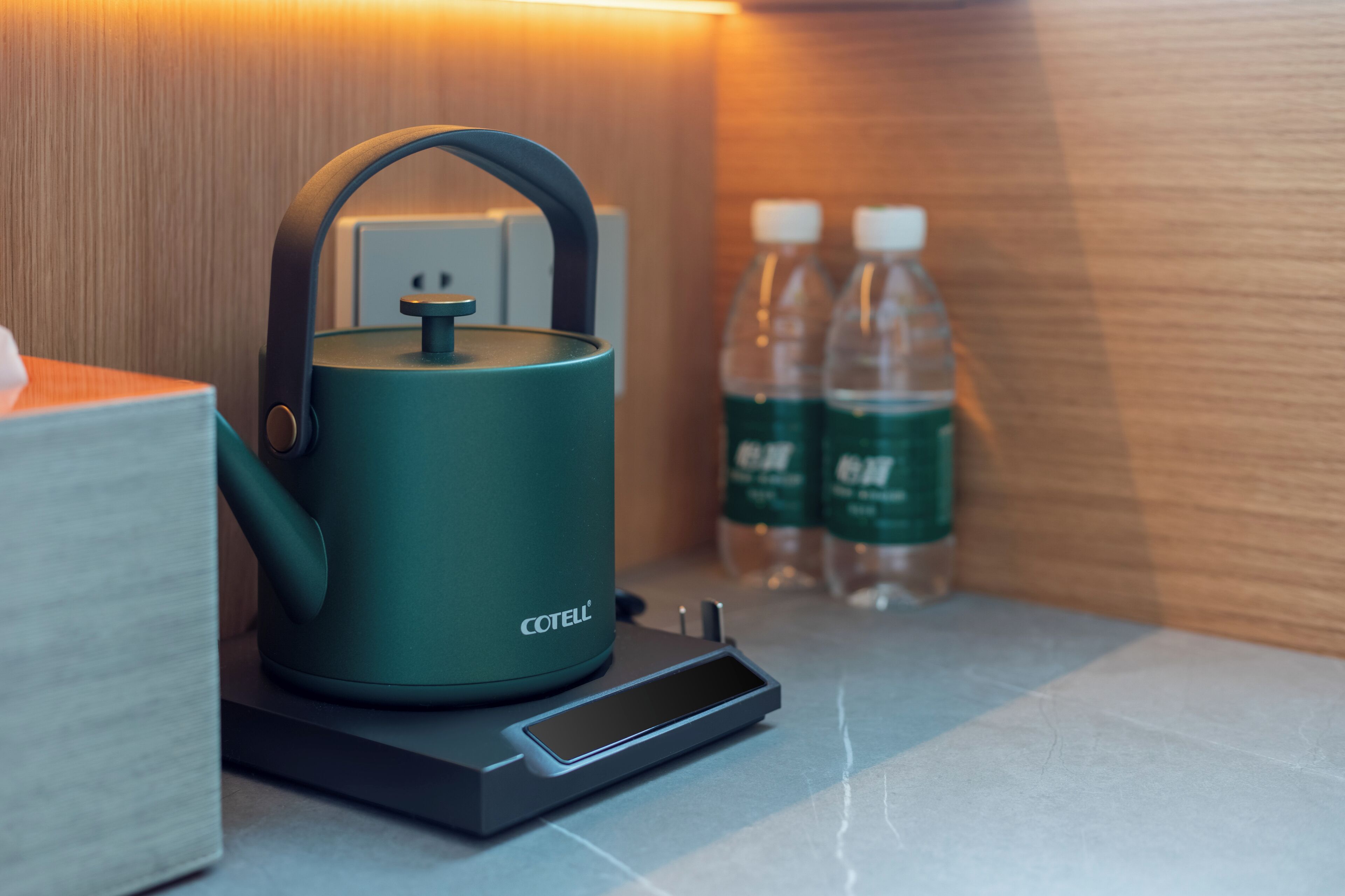 A teal electric kettle on a countertop with a power outlet and two water bottles in the background.