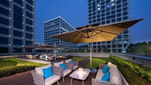 An outdoor seating area with wicker furniture, a large umbrella, and a view of modern buildings. - Pagoda Hotel Hangzhou Zijingang - Near Zhejiang University and Xixi National Wetland Area (Hangzhou)