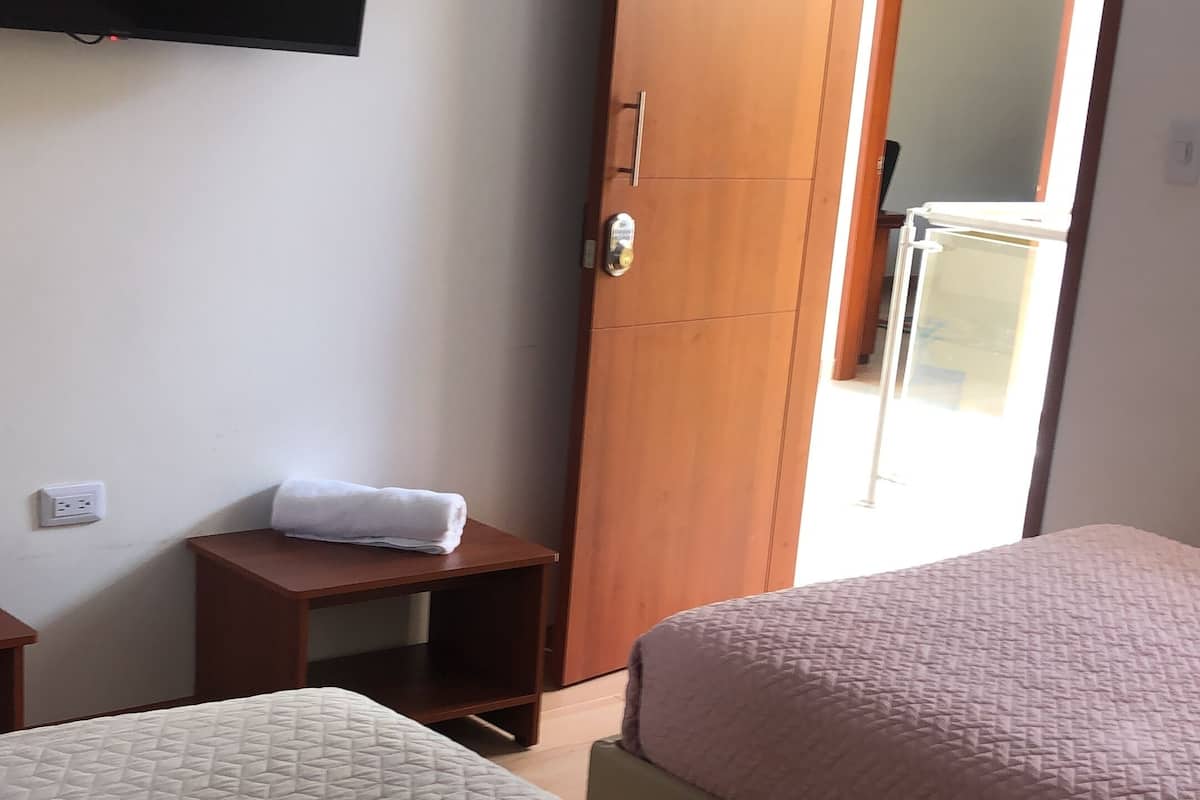 A hotel room with two beds, a TV mounted on the wall, a wooden wardrobe, and a small table with a towel on it.