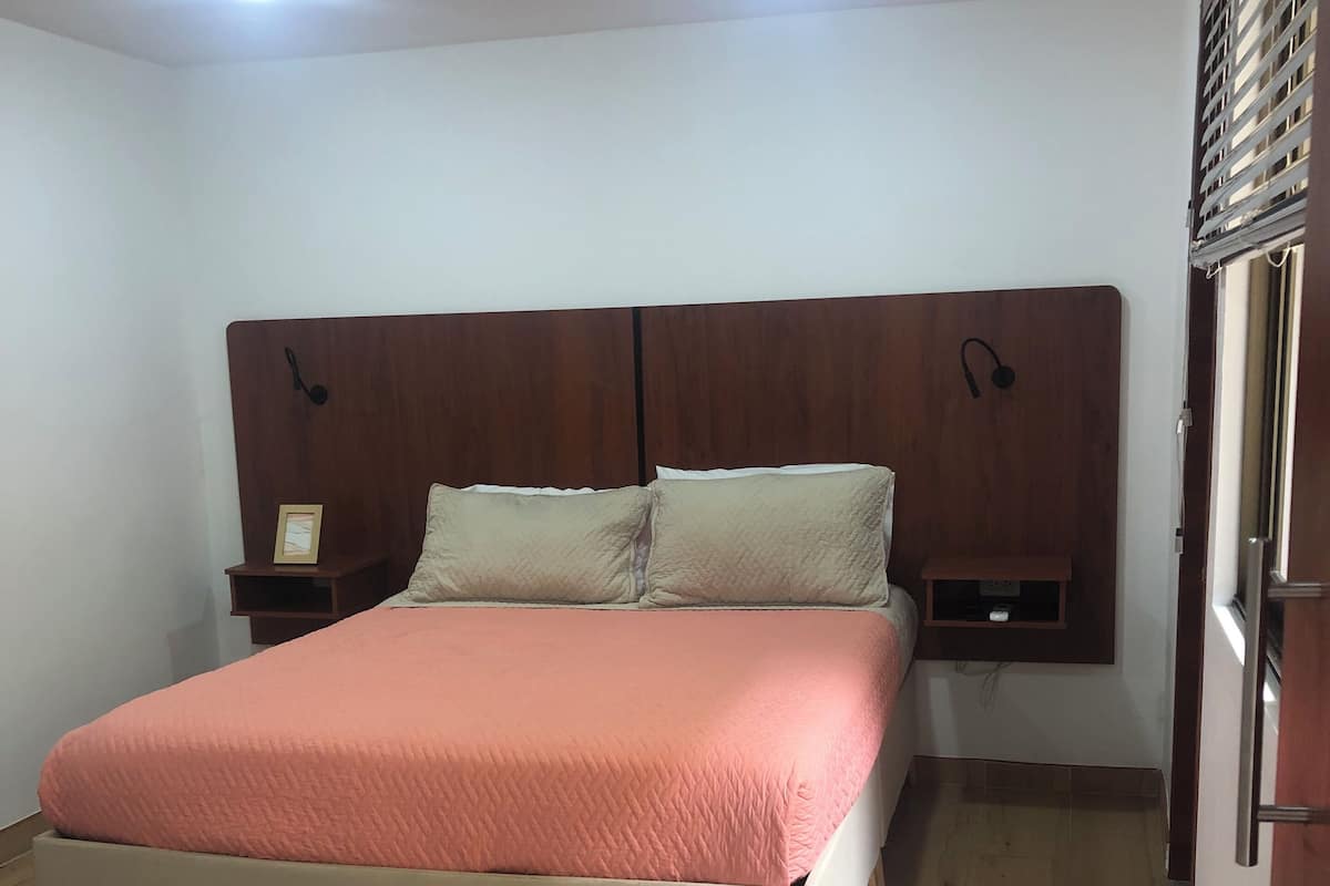 A bedroom with a bed, a colorful rug, wooden flooring, and a headboard with built-in lighting.