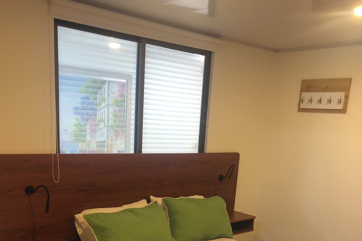A bedroom with a wooden headboard, a bed with green and white bedding, a colorful rug, and a window with blinds.