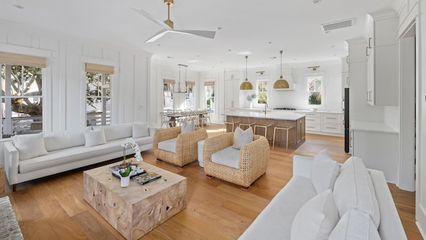 Un vaste salon avec un canapé blanc, des fauteuils en osier et une table basse en bois. On aperçoit au fond une cuisine avec des meubles blancs et un coin repas comprenant une table et des chaises.