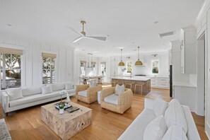 Uma sala de estar espaçosa com um sofá branco, cadeiras de vime e uma mesa de centro de madeira. É visível, ao fundo, uma cozinha com armários brancos e uma área de refeições com mesa e cadeiras.