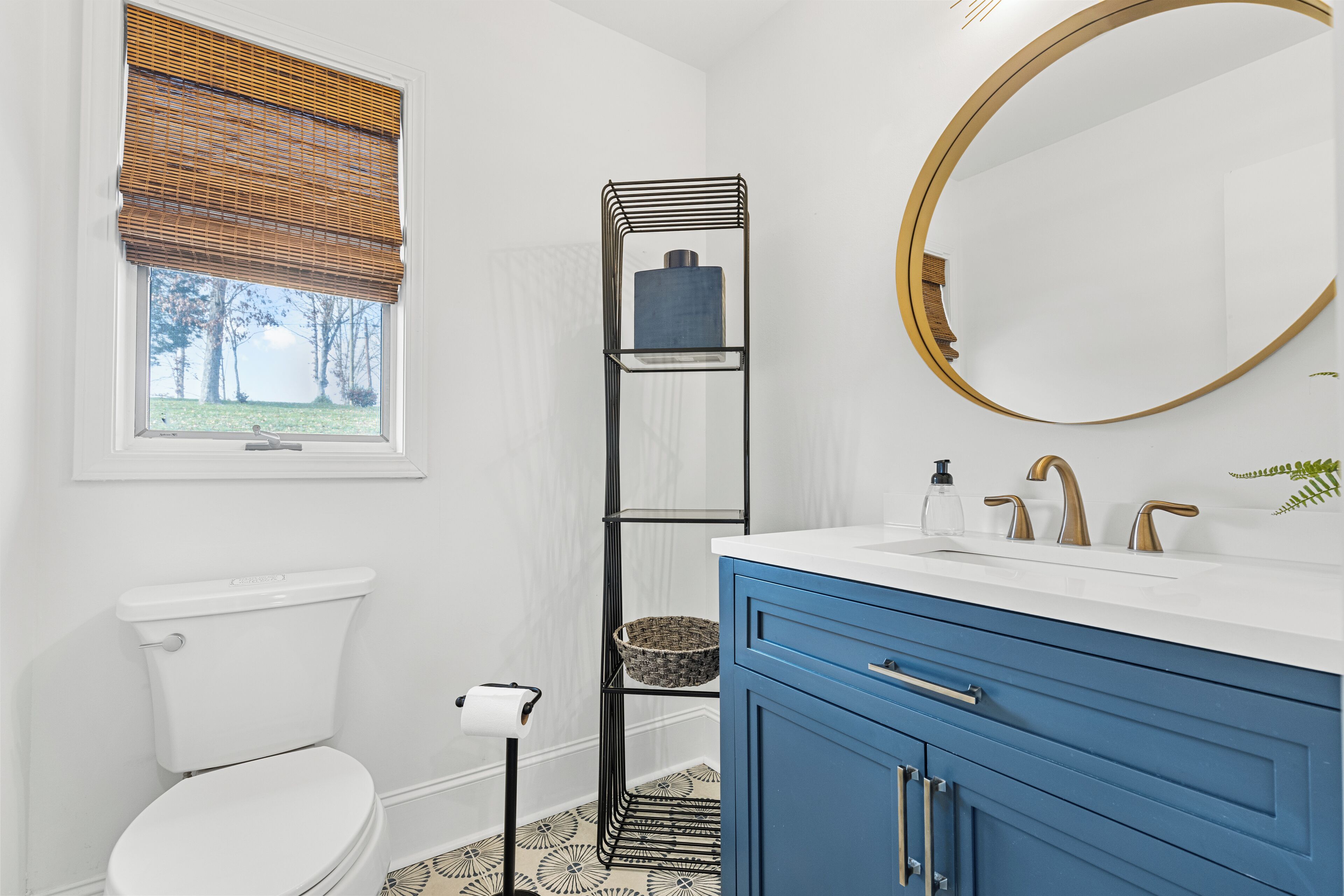 A bathroom with a toilet, a window with a blind, a shelf with a basket, a sink with a mirror, and a blue cabinet.