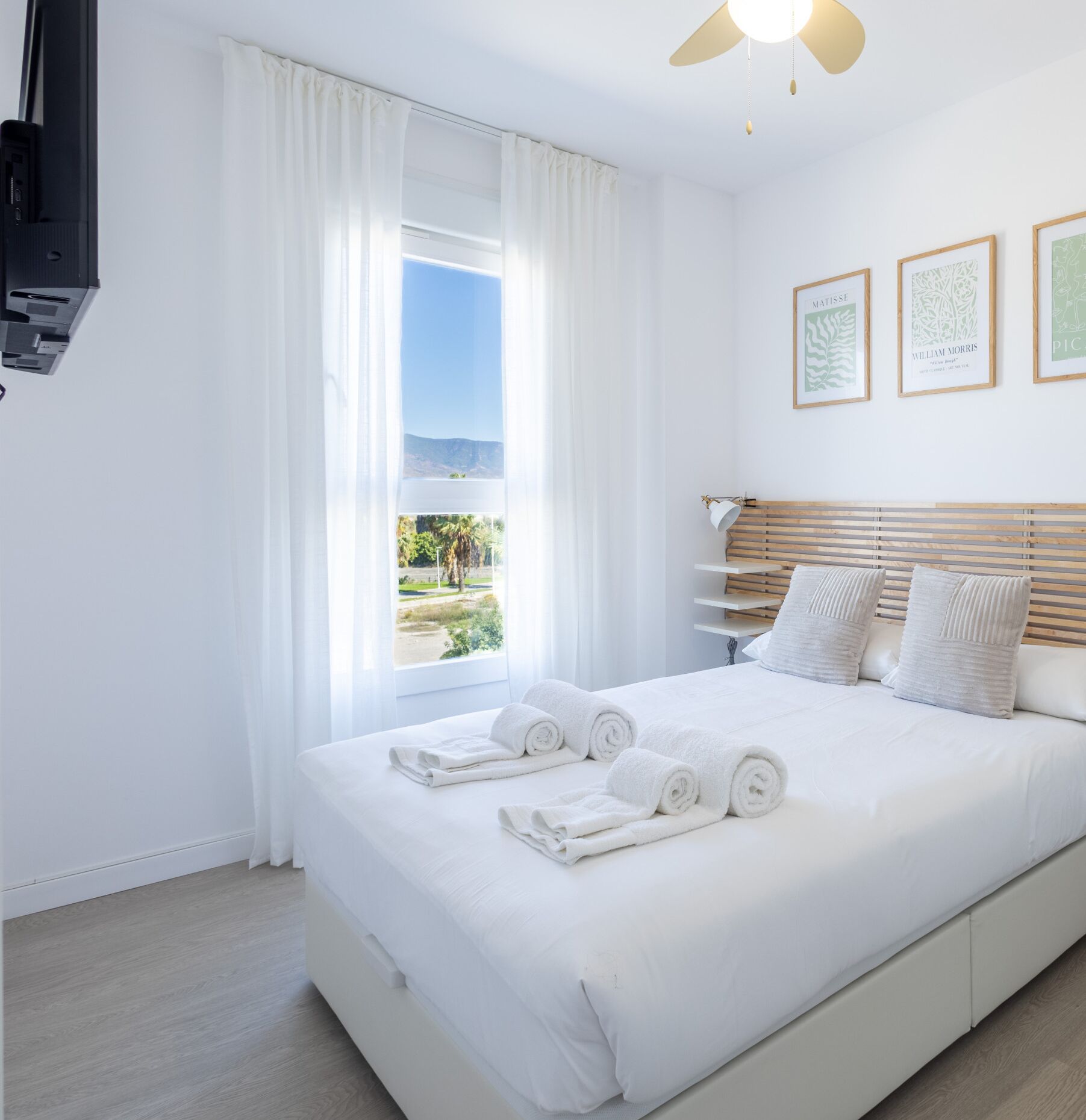 Quarto de hotel com cama, cortinas brancas, ventilador de teto e quadros nas paredes.