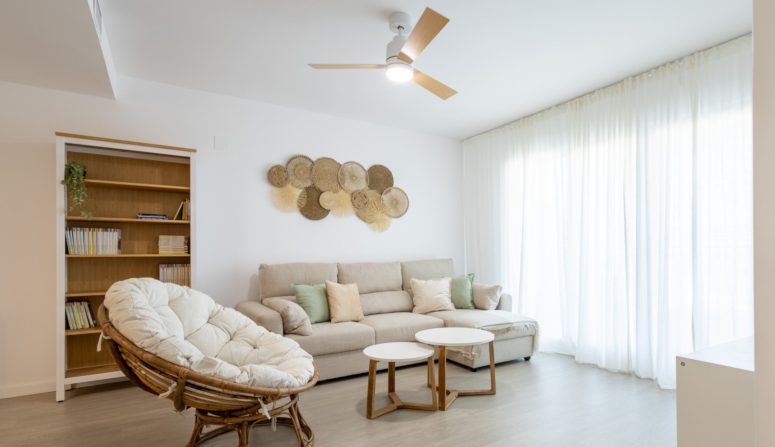 Sala de estar com sofá, duas mesas redondas, uma cadeira de vime, ventilador de teto, estante e uma obra de arte na parede.