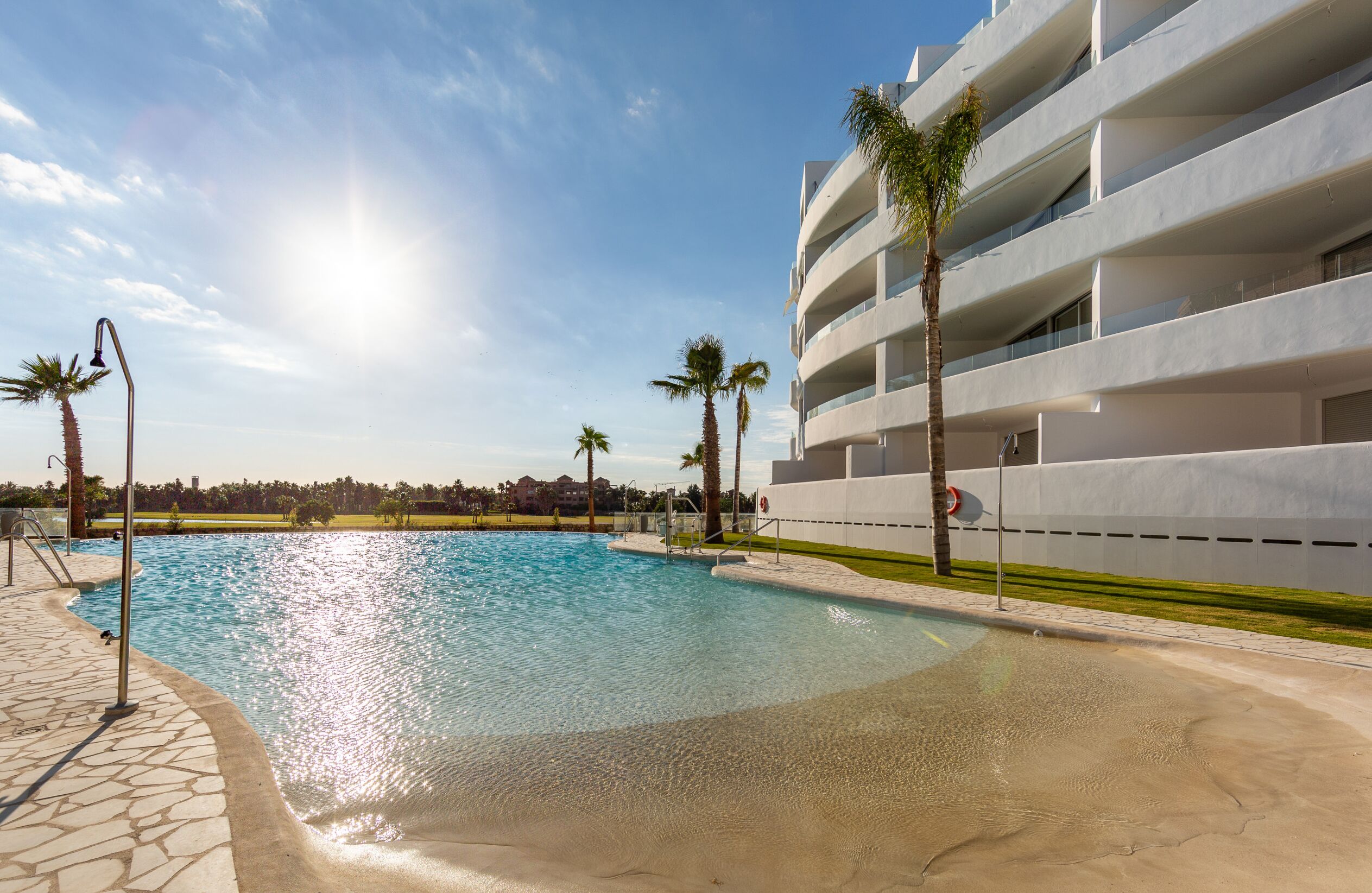 A modern hotel with a swimming pool, palm trees, and a clear sky.