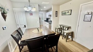 A dining area with a wooden table and chairs, a chandelier, and a kitchen in the background.