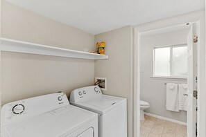 Et vaskerum med to hvide vaskemaskiner, en hylde med en gul æske, samt et badeværelse med toilet og håndklæder.