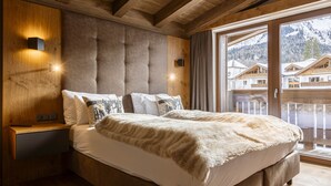 A bedroom with a large bed, wooden headboard, and a view of mountain houses through a glass door. - Beautiful Chalet With Swimming Pool (Ehrwald)