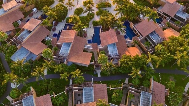 Vue aérienne d’un complexe hôtelier comprenant plusieurs hébergements de type bungalow, une plage et des palmiers.