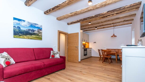 A red sofa with reindeer pillows, a wooden dining table, and a kitchen area with wooden beams and white cabinets.
