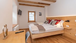 A bedroom with a wooden bed, a TV mounted on the wall, a small window, and a wooden desk with a chair.