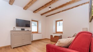 A room with a red sofa, a TV on a cabinet, wooden beams on the ceiling, and windows with blinds.