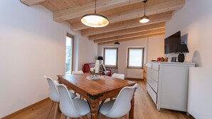 Een eetgedeelte met een houten tafel en witte stoelen, een keuken met een witte koelkast en kasten, en een televisie die aan de muur is bevestigd.