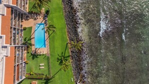Vista aérea de un hotel con piscina, sillones de descanso y un litoral rocoso.